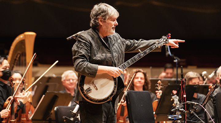 Musician standing on stage, holding a banjo, and pointing while surrounded by orchestra musicians sitting in chairs.